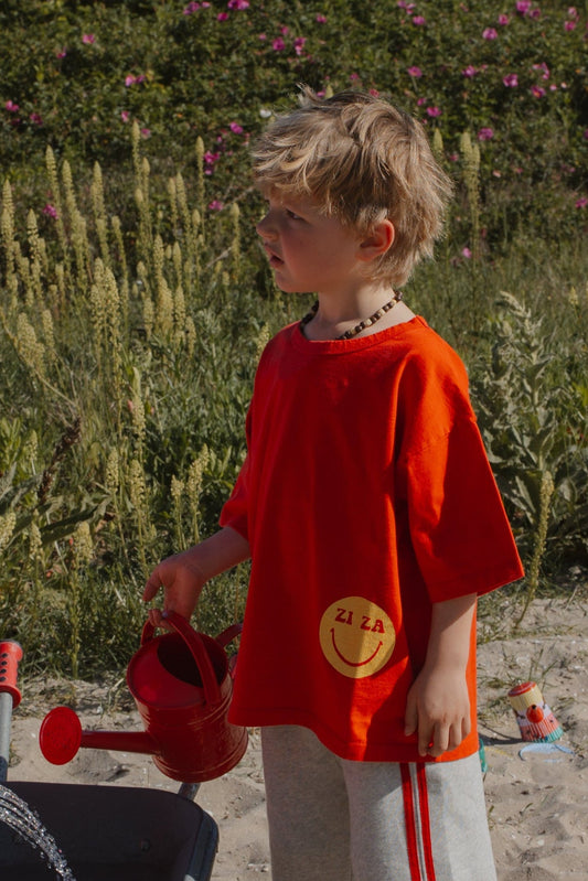 Kid wearing the red ZIZA CLUB Tee SMILE Tomato, an oversized kids t-shirt with a yellow smiley print